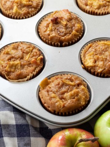 Apple Quinoa Muffins in a muffin tin resting on a checkered dish towel flanked by fresh red and green apples