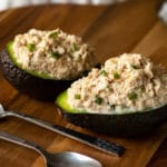 Two avocado halves stuffed with classic tuna salad served on a wooden board with two spoond