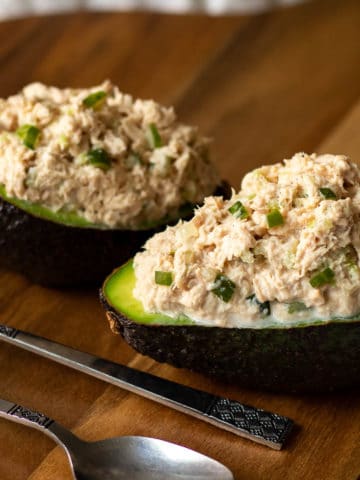 Two avocado halves stuffed with classic tuna salad served on a wooden board with two spoond