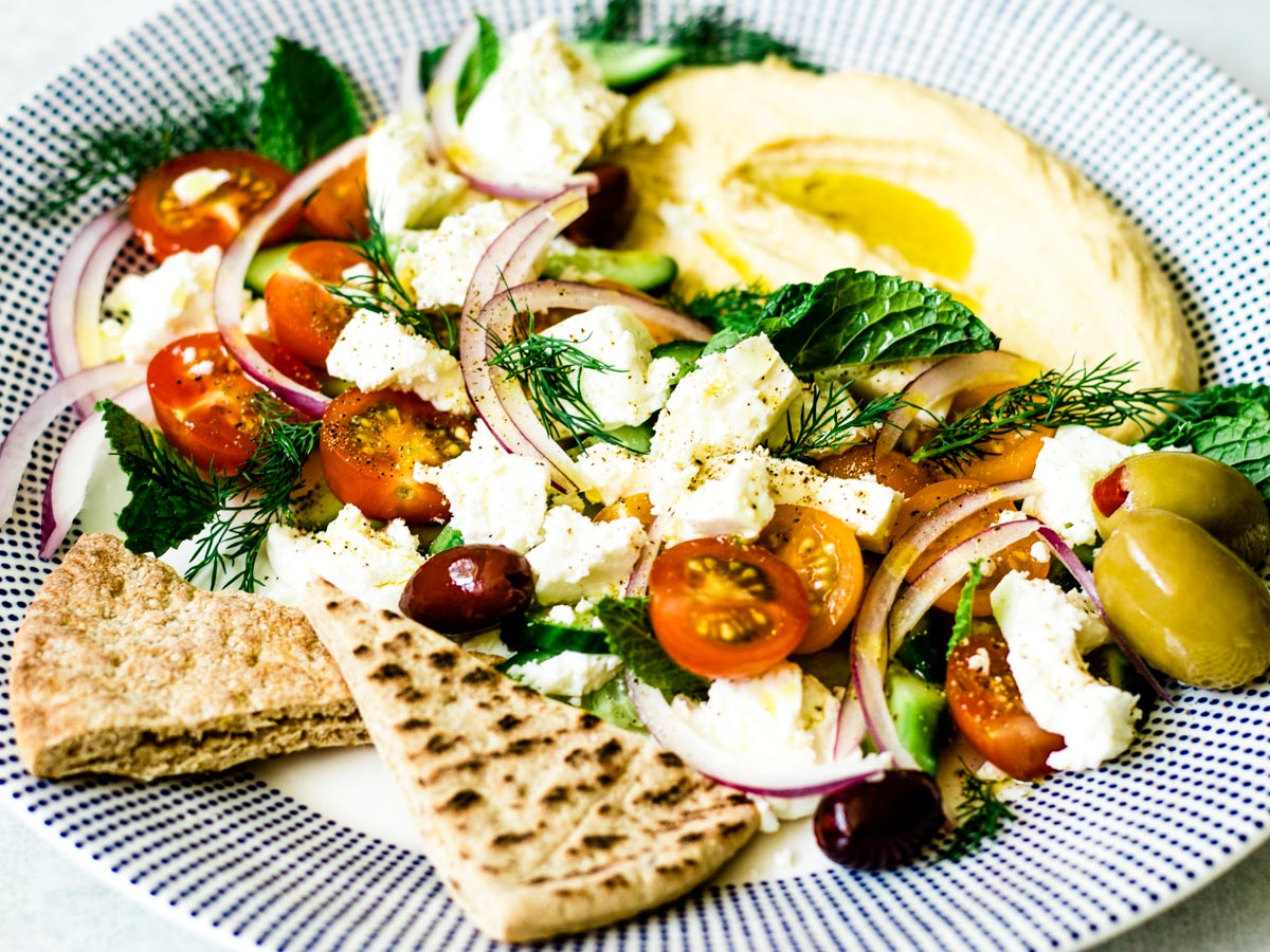Greek salad of tomatoes, cucumbers, mint and dill dressed with olive oil and lemon juice and topped with feta cheese and olives on a platter with pita wedges and hummus