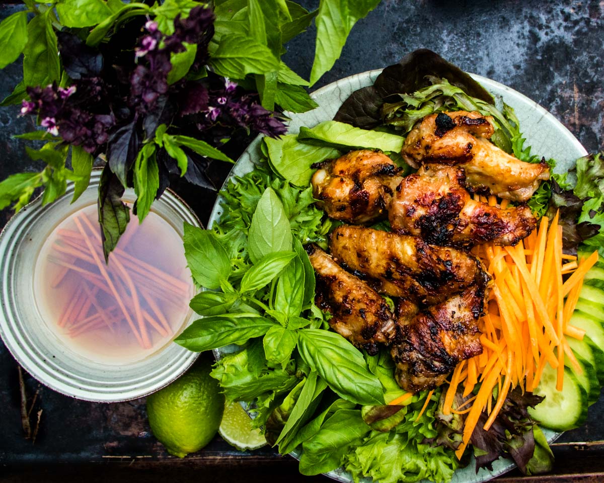 Overhead view of grilled lemongrass chicken wings on a bed of spring greens, basil, mint, cucumbers and shredded carrots with a side of fish sauce and a bouquet of purple and green basil.