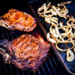 Two seasoned rib-eye steaks on the grill with onion rings