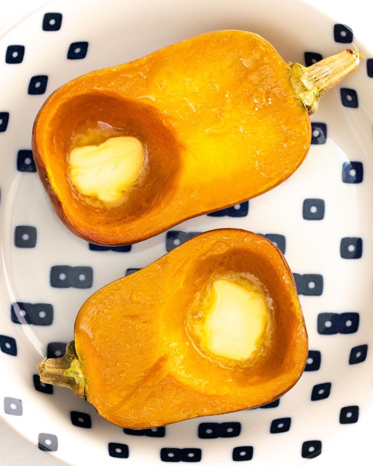 Roasted honeynut squash halves with a pat of melting butter in each cavity served on a white plate decorated with blue squares