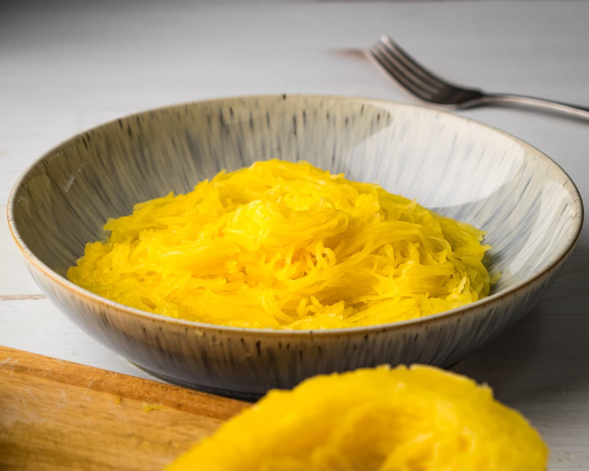 A mound of spaghetti squash noodles in a pasta bowl with a fork and a cutting board with cut spaghetti squash