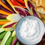 A bowl of fresh keto dill dip surrounded with cucumbers, endive, carrots, bell pepper, cherry tomatoes and pita for dipping