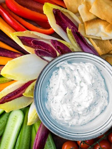 A bowl of fresh keto dill dip surrounded with cucumbers, endive, carrots, bell pepper, cherry tomatoes and pita for dipping