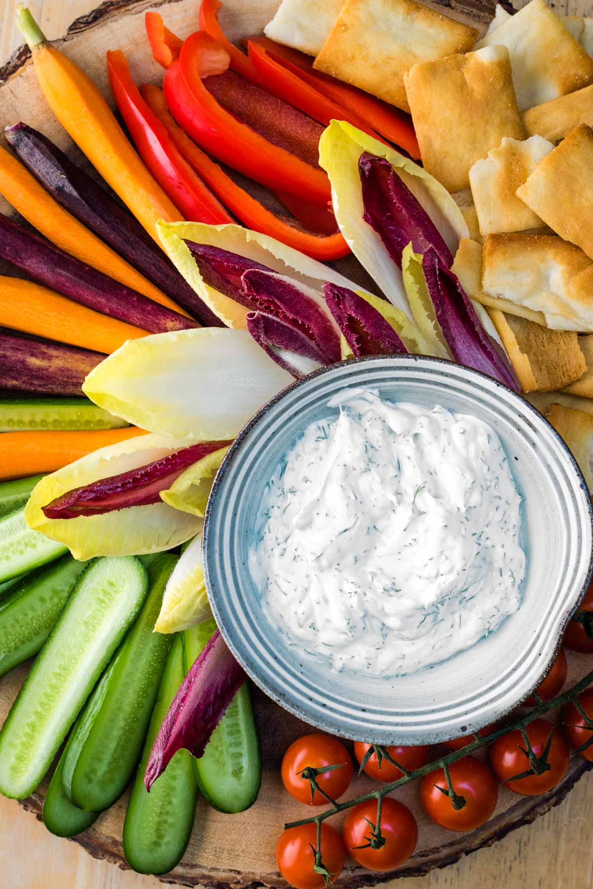 A bowl of fresh keto dill dip surrounded with cucumbers, endive, carrots, bell pepper, cherry tomatoes and pita for dipping