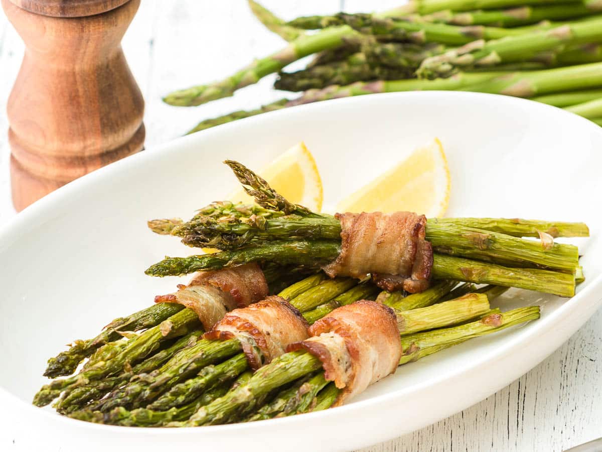 Air fryer bacon wrapped asparagus on a plate with lemon wedges and pepper mill