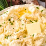 a bowl of cauliflower mashed potatoes with chives and melted butter served on a board with a pepper mill