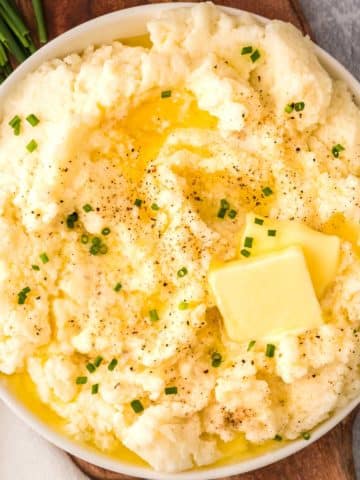 Cauliflower mashed potatoes in a serving bowl with 2 pats of butter salt pepper and chives on a cutting boardoverhead view