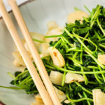Pea shoots plated with chopsticks in forefront resting on the edge of the plate